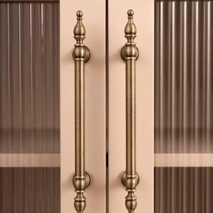 May include: Two antique brass cabinet pulls with a vertical bar design. The pulls have decorative finials at each end and are mounted on a light-colored cabinet door with a ribbed glass panel.