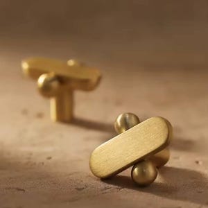 Może przedstawiać: Dwa złote gałki do szafek o nowoczesnym designie. Każda gałka ma prostokątny kształt z zaokrąglonymi końcami, połączony z kulistą podstawą. Gałki wykonane są z błyszczącego metalu.