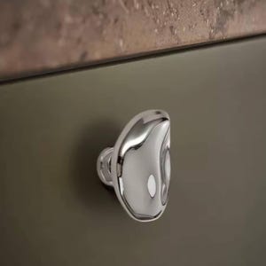 May include: A polished chrome cabinet knob with a unique, organic shape. The knob is mounted on a dark olive-green cabinet door, reflecting light and adding a touch of modern elegance to the design.