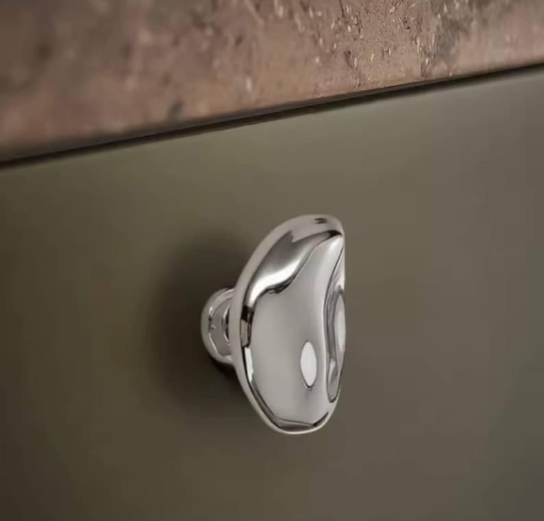 May include: A polished chrome cabinet knob with a unique, organic shape. The knob is mounted on a dark olive-green cabinet door, reflecting light and adding a touch of modern elegance to the design.