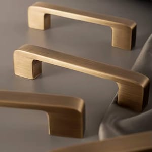 Puede incluir: Primer plano de varios tiradores de armario de color bronce. Los tiradores tienen un dise&ntilde;o rectangular y minimalista con un asa ligeramente curvada. El herraje es de metal y tiene un acabado cepillado. Los tiradores est&aacute;n dispuestos sobre un fondo neutro.