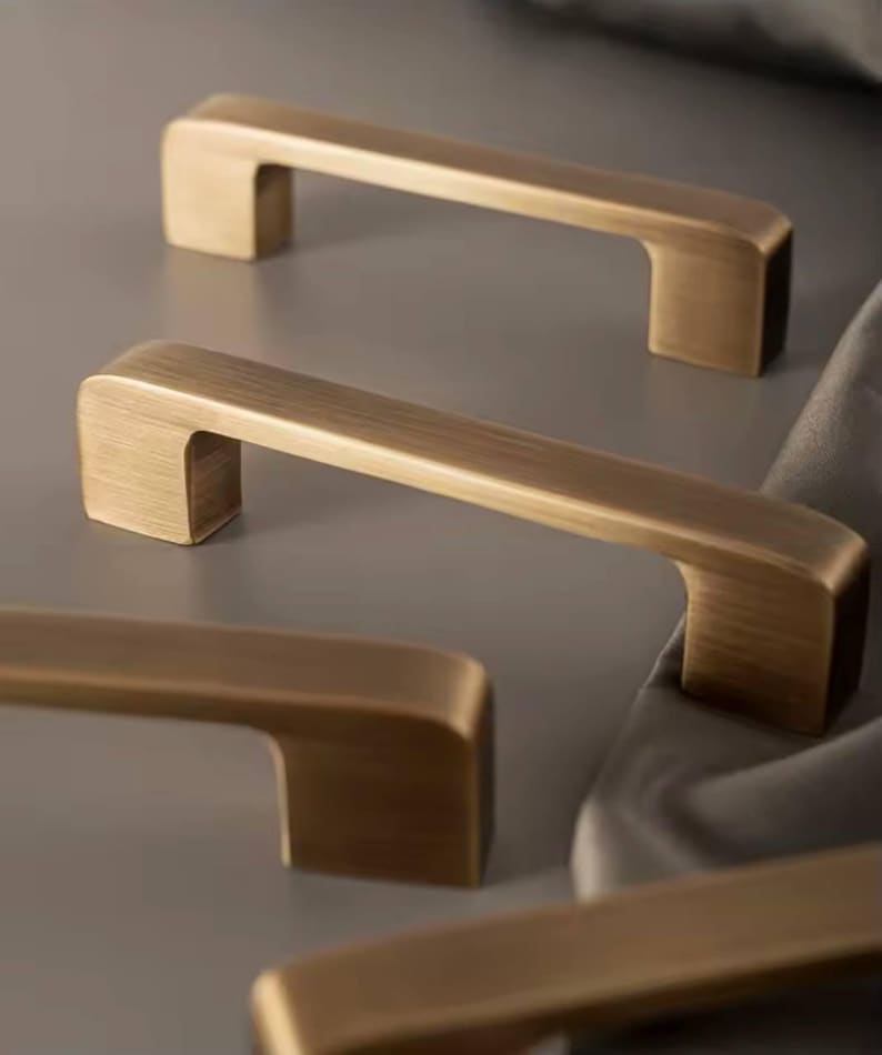 Puede incluir: Primer plano de varios tiradores de armario de color bronce. Los tiradores tienen un dise&ntilde;o rectangular y minimalista con un asa ligeramente curvada. El herraje es de metal y tiene un acabado cepillado. Los tiradores est&aacute;n dispuestos sobre un fondo neutro.
