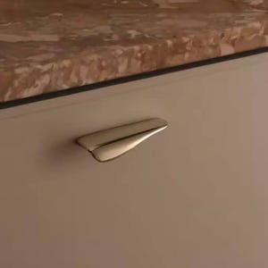 May include: A polished, curved metal cabinet pull with a streamlined design. The handle is mounted on a light beige cabinet door, with a marble countertop visible above. The hardware adds a modern touch to the cabinetry.