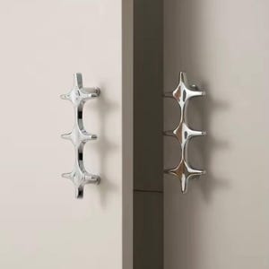 May include: Two chrome cabinet pulls with a starburst design. Each pull has three star-shaped elements connected vertically. The pulls are mounted on a light-colored cabinet door, with a darker cabinet edge visible.