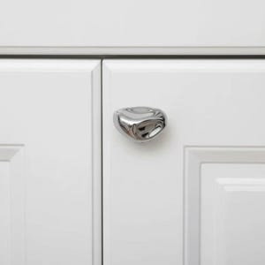 May include: A close-up of a polished chrome cabinet knob with a unique, organic shape. The knob is mounted on a white cabinet door, with a second door visible. The hardware adds a modern touch to the cabinetry.