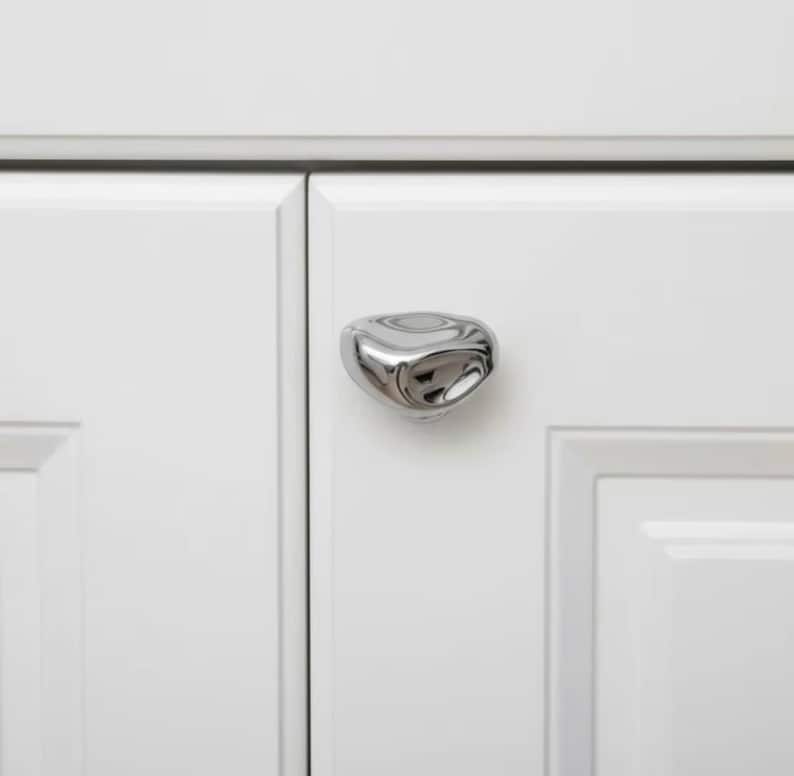May include: A close-up of a polished chrome cabinet knob with a unique, organic shape. The knob is mounted on a white cabinet door, with a second door visible. The hardware adds a modern touch to the cabinetry.