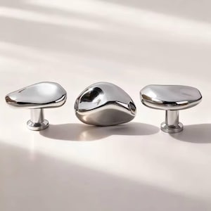 May include: Three polished chrome cabinet knobs with organic, pebble-like shapes. Each knob has a rounded top and a cylindrical base. The knobs are arranged in a row against a neutral background, reflecting light.