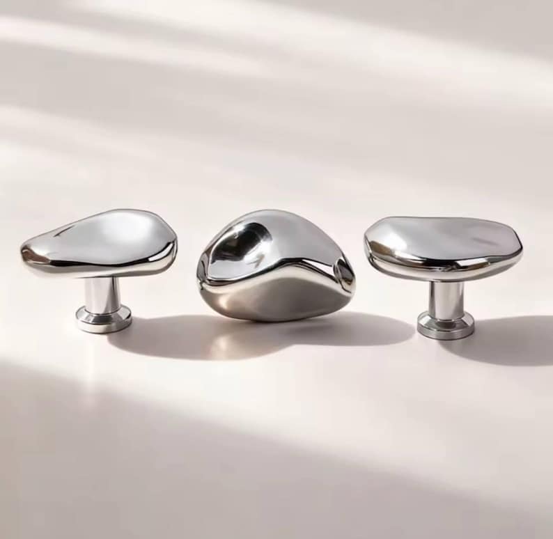 May include: Three polished chrome cabinet knobs with organic, pebble-like shapes. Each knob has a rounded top and a cylindrical base. The knobs are arranged in a row against a neutral background, reflecting light.