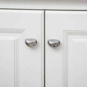 May include: Close-up of a white cabinet with two silver, organic-shaped cabinet knobs. The knobs have a smooth, reflective surface and are mounted on the cabinet doors. The cabinet doors are white with a recessed panel design.