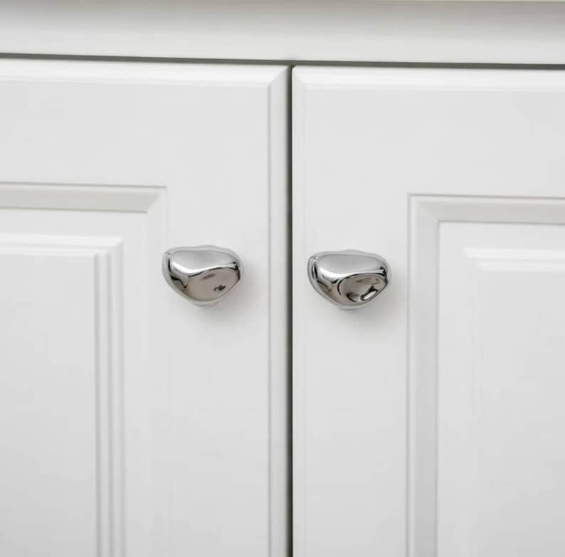 May include: Close-up of a white cabinet with two silver, organic-shaped cabinet knobs. The knobs have a smooth, reflective surface and are mounted on the cabinet doors. The cabinet doors are white with a recessed panel design.
