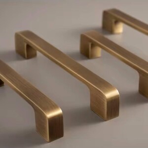 Puede incluir: Cuatro tiradores de armario de lat&oacute;n antiguo dispuestos sobre una superficie neutra. Cada tirador tiene una barra rectangular con bordes curvos y una base rectangular. Los tiradores son de un color met&aacute;lico c&aacute;lido, lo que sugiere una est&eacute;tica vintage o industrial.