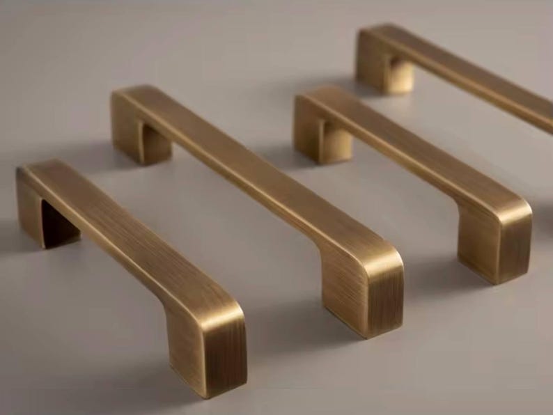 Puede incluir: Cuatro tiradores de armario de lat&oacute;n antiguo dispuestos sobre una superficie neutra. Cada tirador tiene una barra rectangular con bordes curvos y una base rectangular. Los tiradores son de un color met&aacute;lico c&aacute;lido, lo que sugiere una est&eacute;tica vintage o industrial.
