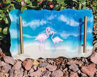 Puede incluir: Una bandeja rectangular para servir con un diseño de olas de mar azul y blanco y dos flamencos rosas. La bandeja tiene asas de metal dorado a cada lado.