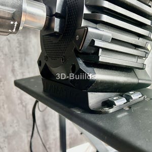 Op de afbeelding: Close-up van een zwarte gaming stuurwielbasis met de tekst "3D-Buiilds" en "FANATEC" zichtbaar. De basis is gemonteerd op een zwart oppervlak en heeft diverse mechanische componenten en montagehardware.