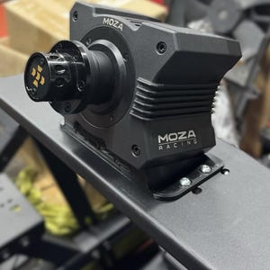 May include: A black MOZA Racing direct drive wheel base. The device is mounted on a black metal surface. The wheel base has the MOZA Racing logo and a gold and black emblem on the front.