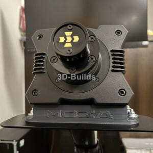 Może przedstawiać: Czarna podstawa kierownicy wyścigowej MOZA ze złotym wzorem strzałki na środkowej nasadce. Podstawa ma prostokątny kształt z wieloma śrubami i napisem "3D-Builds". Podstawa jest zamontowana na czarnej metalowej płycie.