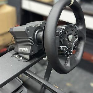 Op de afbeelding: Een zwart MOZA Racing sim race stuur en basis gemonteerd op een zwart metalen frame. Het stuur is zwart met witte accenten en meerdere knoppen. De basis heeft het MOZA Racing logo.