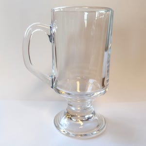 May include: A clear glass mug with a handle, a pedestal base, and a cylindrical body. The mug is empty and appears to be made of transparent glass. The handle is curved and attached to the side of the mug.