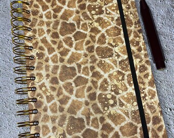 May include: A brown and tan spiral bound notebook with a giraffe print cover. The notebook has a black elastic band closure and a gold metal spiral binding. The notebook has a gold foil initial "C" on the front cover.