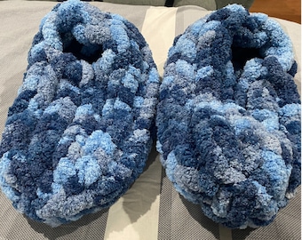 May include: A pair of cozy, handmade slippers in shades of blue. The slippers are made of a thick, plush yarn, with a textured, looped appearance. The color palette includes light, medium, and dark blues, creating a marbled effect.