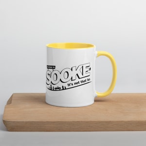 Puede incluir: Taza de cerámica blanca con borde amarillo. La taza tiene un gráfico negro que dice "Bienvenido a Sooke. No está tan lejos."