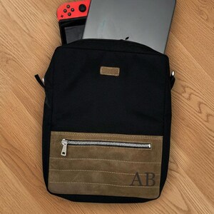 May include: Black messenger bag with a brown suede-like front pocket and a small leather patch. The bag contains a grey tablet and a red and black game console. The initials "AB" are visible on the front.
