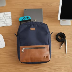 May include: A navy blue backpack with a brown leather-like front pocket and a silver zipper. The initials "AB" are visible on the pocket. A tablet and a teal game console are partially visible inside the backpack.