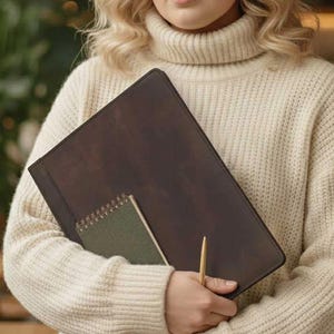 Può includere: Una cartella in pelle marrone scuro contiene un piccolo taccuino verde a spirale e una penna dorata. La cartella è rettangolare e sembra chiusa. Il taccuino è piccolo e rettangolare. La penna è sottile e dorata.