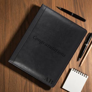 Op de afbeelding: Zwart leren portfolio met het woord "Gefeliciteerd" en initialen "AB" in reliëf op de omslag. De portfolio ligt op een houten oppervlak, vergezeld van twee zwarte pennen, een potlood en een klein notitieblok.