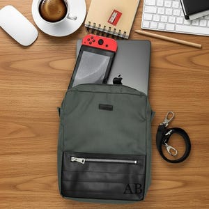 May include: A grey backpack with a black, leather-look base, silver zip, and initials "AB." Inside is a laptop and a red and black games console. A black strap with silver fittings lies beside the bag.