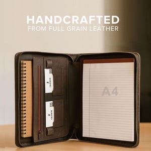 May include: Open, dark brown leather portfolio with a zipper closure. The portfolio contains a spiral-bound notebook, a notepad, and card slots. The text "HANDCRAFTED FROM FULL GRAIN LEATHER" is displayed at the top. The notepad has the text "A4" printed on it.