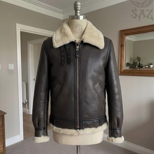 May include: A dark brown leather jacket featuring a cream-colored shearling collar and trim at the cuffs and hem. The jacket includes a central metal zipper, two side pockets, and two adjustable buckle straps located on the upper chest area.