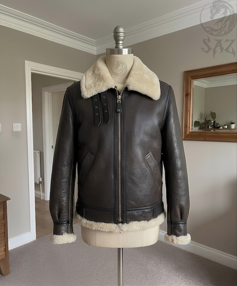 May include: A dark brown leather jacket featuring a cream-colored shearling collar and trim at the cuffs and hem. The jacket includes a central metal zipper, two side pockets, and two adjustable buckle straps located on the upper chest area.