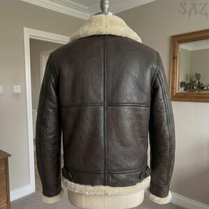 May include: A dark brown leather jacket featuring a thick cream-colored shearling collar, cuffs, and hem. The jacket has visible vertical and horizontal seam detailing across the back, along with adjustable side buckle straps at the waist.