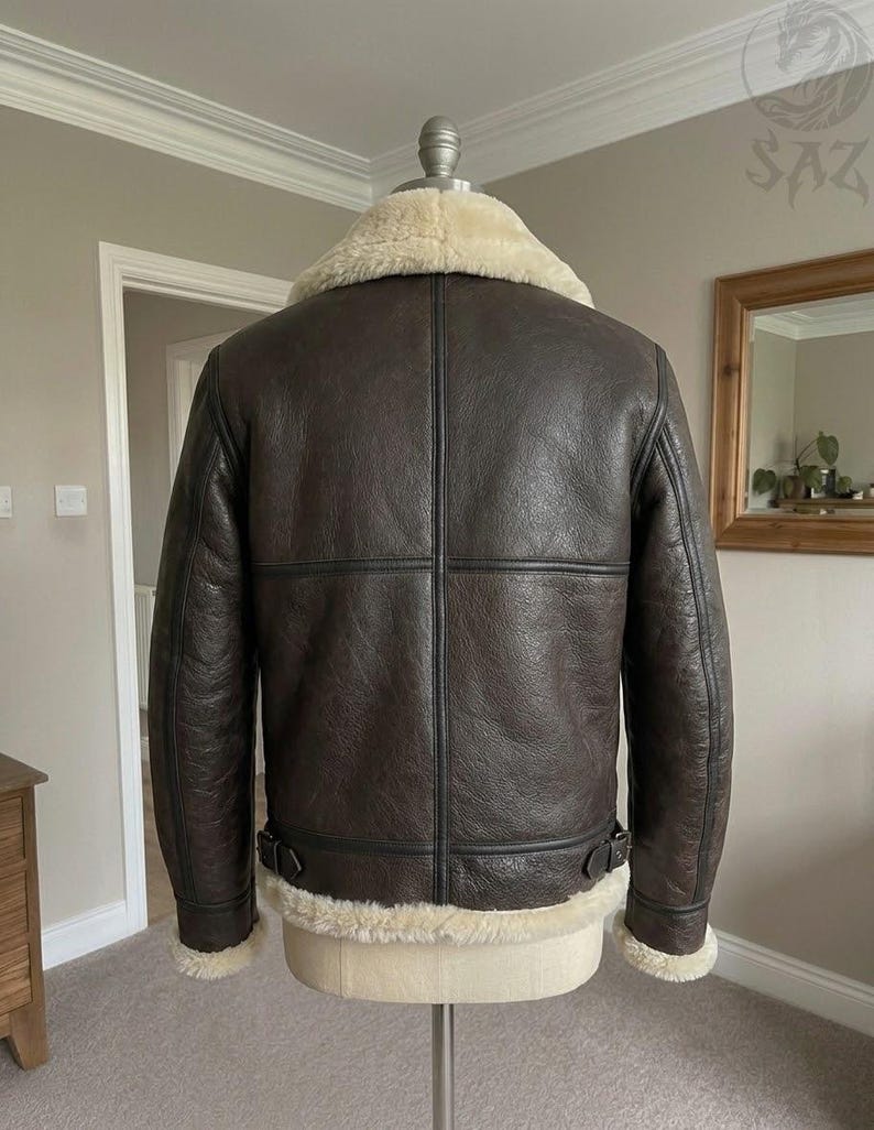 May include: A dark brown leather jacket featuring a thick cream-colored shearling collar, cuffs, and hem. The jacket has visible vertical and horizontal seam detailing across the back, along with adjustable side buckle straps at the waist.