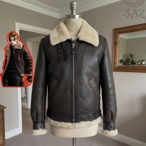 May include: A dark brown leather bomber jacket featuring a cream-colored shearling collar, cuffs, and hem. The jacket includes a central metal zipper, two black buckled throat latch straps on the collar, and long sleeves on a mannequin torso.