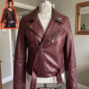 Pode incluir: Um casaco de motociclista em pele vermelho-escuro com gola larga, fecho de correr frontal assimétrico e um cinto com fivela na cintura. O casaco inclui um bolso no peito com fecho, dois bolsos inferiores com fecho e um pequeno alfinete.
