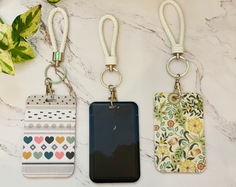 Puede incluir: Tres llaveros con diferentes porta tarjetas estampados. El primer llavero tiene un porta tarjetas estampado en blanco, gris y rosa con corazones y formas geométricas. El segundo llavero tiene un porta tarjetas negro con una luna creciente blanca. El tercer llavero tiene un porta tarjetas blanco con un estampado floral en tonos verdes, amarillos y rojos.