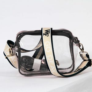 May include: Clear plastic crossbody bag with a black and cream adjustable strap. The strap has the text "WF GO DEAC" printed on it.