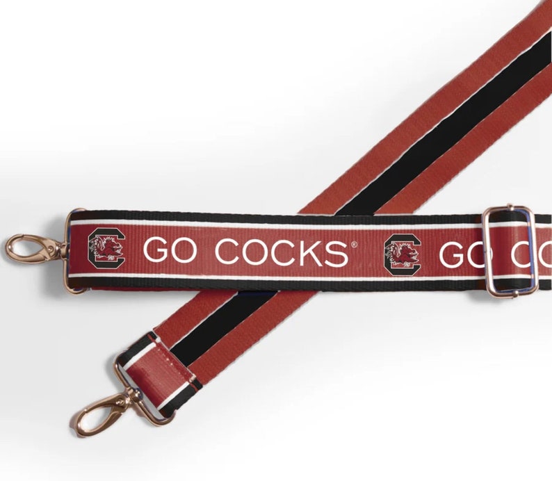 May include: A red and black shoulder strap with the text "GO COCKS" in white. The strap features a repeating pattern of the team logo and is secured with gold-coloured metal clasps.