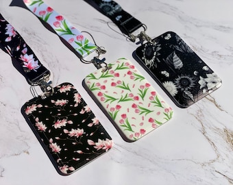 Puede incluir: Tres portagajetas diferentes con diseños florales y cordones a juego. Los portagajetas son blancos con flores rosas, negros con flores rosas y negros con flores blancas. Los cordones son blancos con flores rosas, negros con flores rosas y negros con flores blancas.