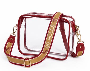 May include: Clear crossbody bag with red trim and a gold and red strap. The strap features the text "GO NOLES" in white. The bag has gold-colored hardware and a rectangular shape. The bag is designed to be transparent.