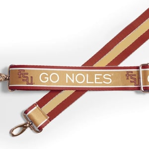 Puede incluir: Una correa de bolso granate y dorada con el texto "GO NOLES" en blanco. La correa presenta cierres metálicos dorados y una franja dorada en el centro. También es visible el logotipo de los Florida State Seminoles.