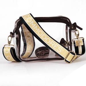 May include: Clear plastic crossbody bag with a brown trim and a gold chain strap. The bag has a black and white striped strap and a tan strap with the text "Commodores" printed in white.