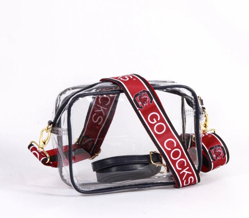 May include: Transparent rectangular crossbody bag with black trim and gold-coloured hardware. The bag has a wide, detachable strap with the text "GO COCKS" in white on a maroon background. A smaller black strap is also included.
