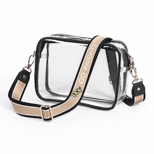May include: Clear crossbody bag with a black trim and a gold and black adjustable strap. The strap features the text "UCF GO KNIGHTS". The bag has gold-colored hardware and a rectangular shape.