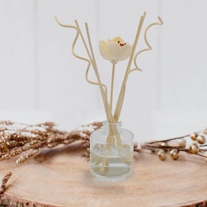 May include: A clear glass diffuser bottle with reeds and a cream-colored flower. The bottle sits on a wooden slice, with gold-colored decorative branches on either side. The background is white.