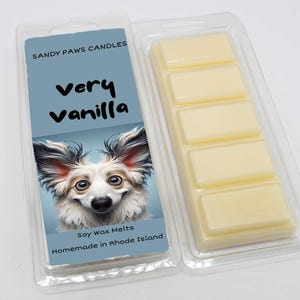 May include: A clear plastic package containing vanilla-scented soy wax melts. The label features a smiling dog illustration and the text "Very Vanilla" and "Sandy Paws Candles". The wax melts are rectangular and light yellow.