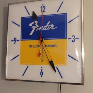 May include: A white square clock with a silver frame. The clock face is divided into two sections, one blue and one yellow. The blue section has the word "Fender" in white lettering. The yellow section has the text "Fine Electric Instruments". The clock hands are black and red.