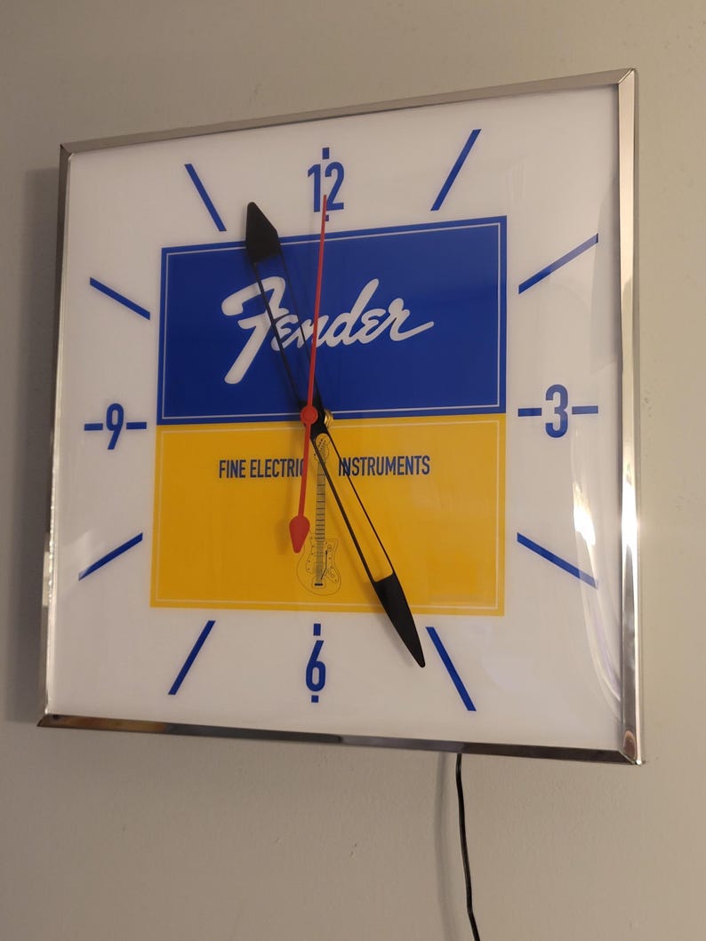 May include: A white square clock with a silver frame. The clock face is divided into two sections, one blue and one yellow. The blue section has the word "Fender" in white lettering. The yellow section has the text "Fine Electric Instruments". The clock hands are black and red.